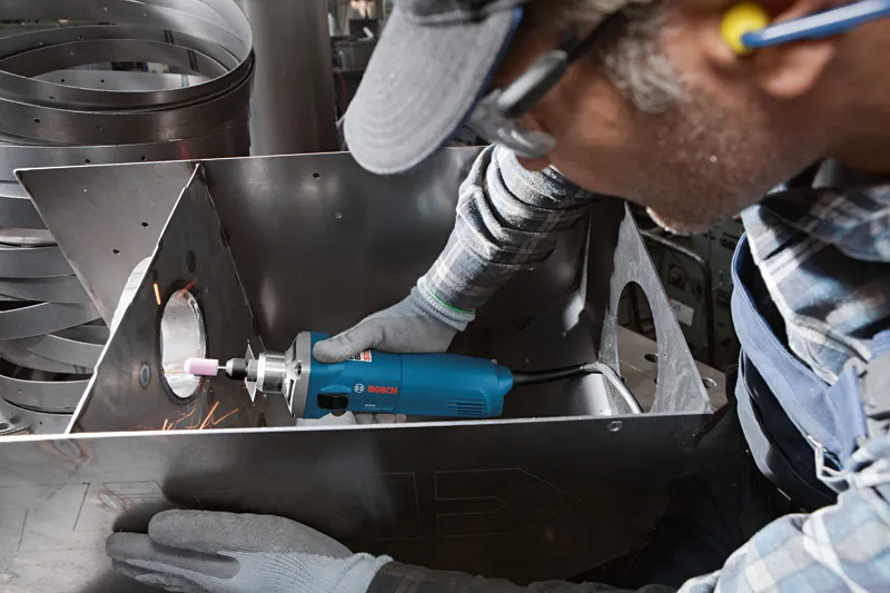 A person wearing safety equipment uses a straight grinder to polish metal inside a duct.